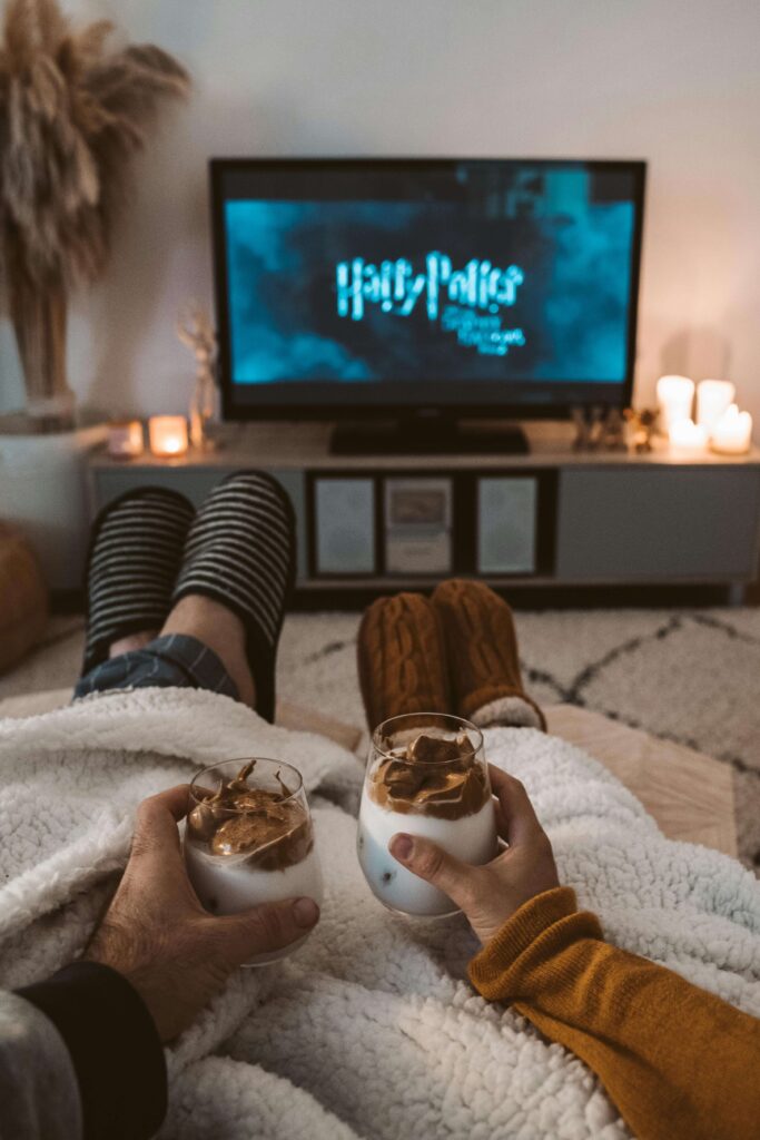 Visão em primeira pessoa dos pés de um casal relaxando sob um cobertor branco, cada um segurando uma bebida quente enquanto assistem filmes para casal, representando uma noite de filme de sucesso.